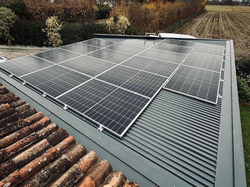 Moduli fotovoltaici su copertura in lamiera grigia, con campi e alberi sullo sfondo